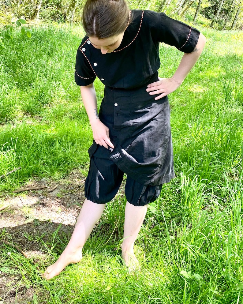 May include: A black dress with a contrasting orange stitch detail. The dress has a button-up front and a gathered skirt. The model is wearing the dress with a pair of black bloomers.