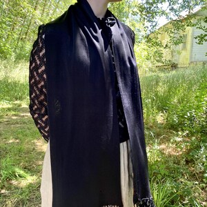 Puede incluir: Una bufanda negra con flecos. La bufanda est&aacute; drapeada sobre una blusa negra con mangas de encaje. El fondo presenta un entorno natural al aire libre con &aacute;rboles y un edificio.