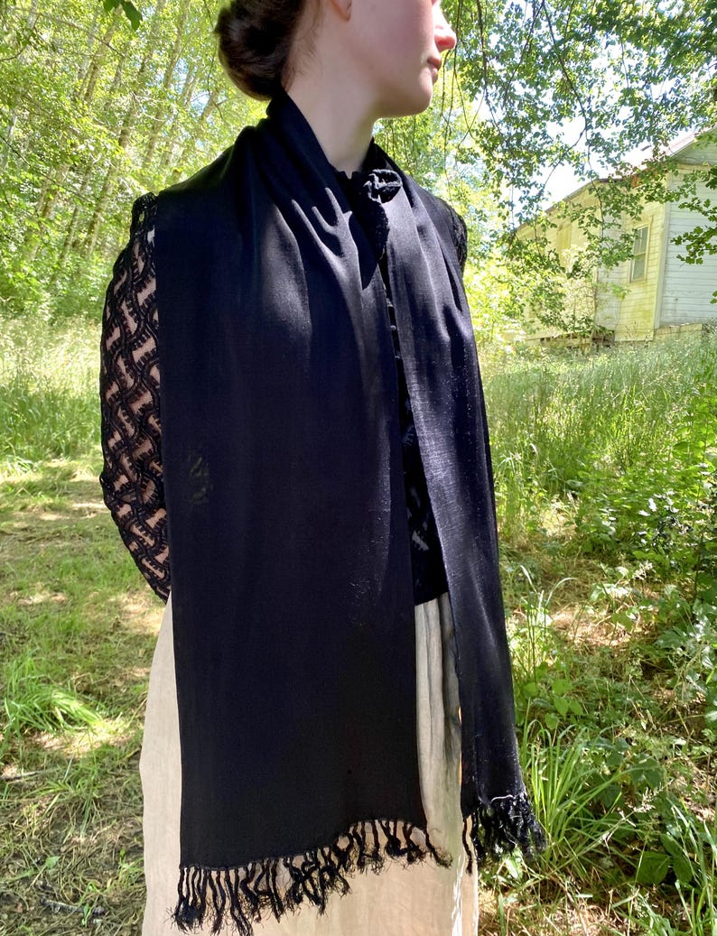 Puede incluir: Una bufanda negra con flecos. La bufanda est&aacute; drapeada sobre una blusa negra con mangas de encaje. El fondo presenta un entorno natural al aire libre con &aacute;rboles y un edificio.