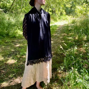 Puede incluir: Un chal negro con flecos se lleva sobre un vestido beige. El chal tiene mangas de encaje y un cierre decorativo. La persona est&aacute; de pie en un camino rodeado de &aacute;rboles y hierba.