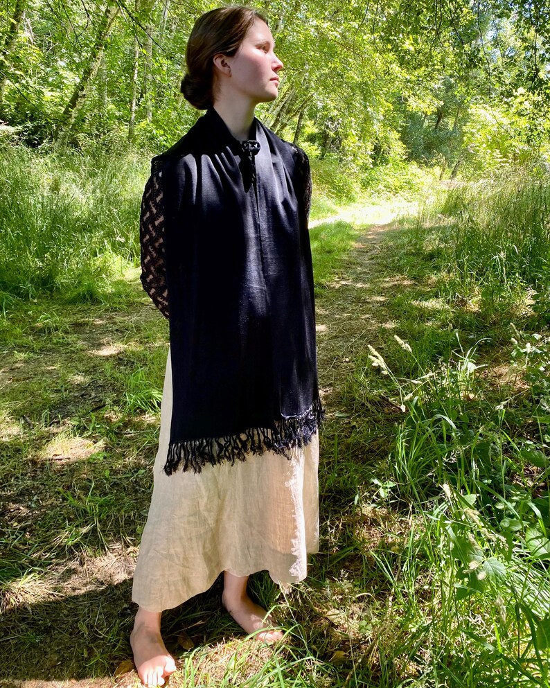 Puede incluir: Un chal negro con flecos se lleva sobre un vestido beige. El chal tiene mangas de encaje y un cierre decorativo. La persona est&aacute; de pie en un camino rodeado de &aacute;rboles y hierba.
