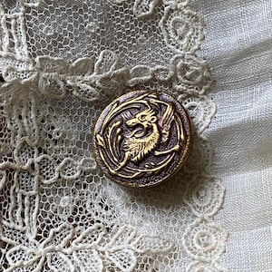 Puede incluir: Un botón redondo y antiguo con un diseño detallado de dragón en dorado sobre un fondo marrón oscuro. El botón se encuentra sobre un fondo de encaje blanco delicado, creando una estética vintage.