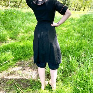 May include: A black dress with a short sleeve and a contrasting red trim around the neckline and sleeves. The dress has a wrap style and a skirt that falls to the knees. The dress is worn by a person standing in a grassy field.