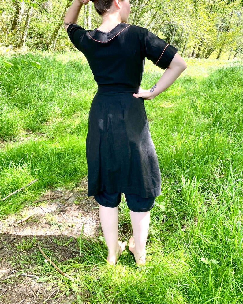 May include: A black dress with a short sleeve and a contrasting red trim around the neckline and sleeves. The dress has a wrap style and a skirt that falls to the knees. The dress is worn by a person standing in a grassy field.