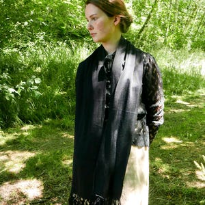 Puede incluir: Una bufanda negra con flecos est&aacute; drapeada sobre los hombros de una persona. La bufanda est&aacute; hecha de un material ligero y cuelga por delante. La persona lleva un top negro y una falda larga color crema. El fondo es un bosque verde.