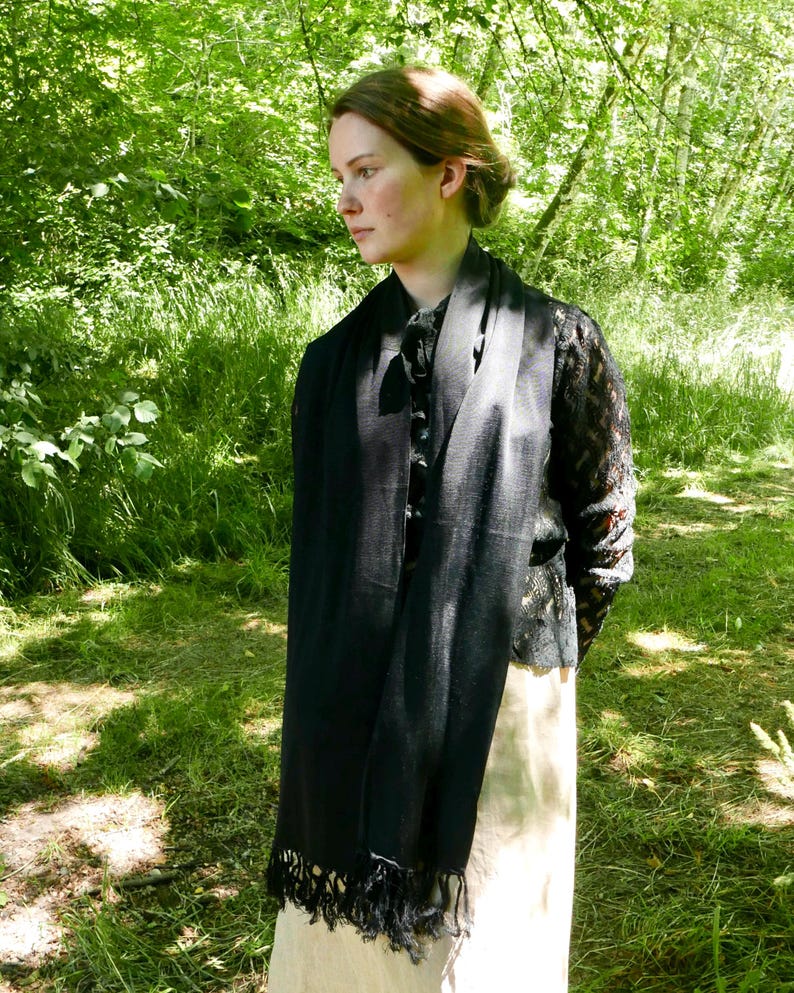 Puede incluir: Una bufanda negra con flecos est&aacute; drapeada sobre los hombros de una persona. La bufanda est&aacute; hecha de un material ligero y cuelga por delante. La persona lleva un top negro y una falda larga color crema. El fondo es un bosque verde.