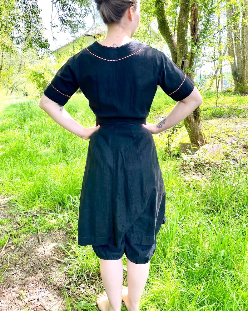 May include: A black linen dress with a short sleeve and a contrasting red stitching detail around the neckline and sleeves. The dress has a wrap style and a gathered skirt.