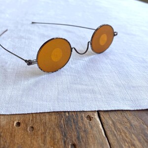 May include: Antique-style spectacles featuring thin, dark metal wire frames and circular, amber-tinted lenses. The glasses have a simple bridge and slender temples that curve outward, showcasing a weathered, aged finish on the metal rims.