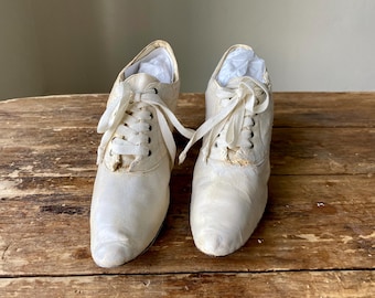 May include: A pair of antique cream-coloured lace-up shoes. The shoes have a pointed toe and a small heel. The laces are long and tied in a bow. The shoes appear to be made of a soft, possibly satin-like material. The shoes are placed on a wooden surface.