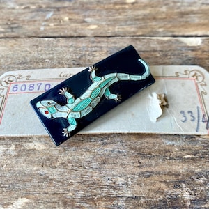 May include: A vintage black enamel brooch featuring a detailed turquoise and white lizard design with a red eye. The lizard is set against a rectangular black background. The brooch is resting on a vintage tag.