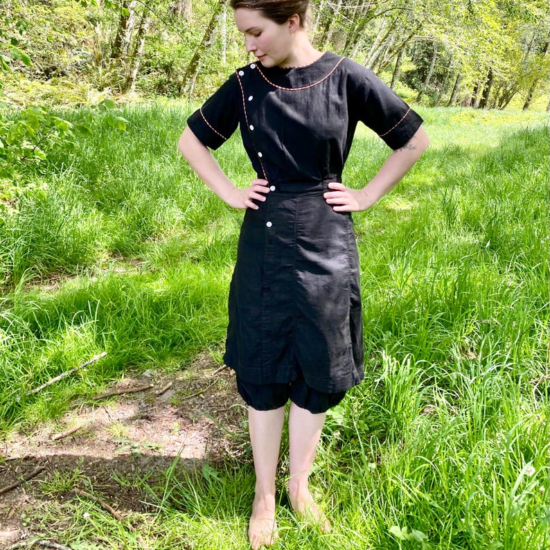 May include: A black dress with a skirt and a top, featuring a button-up closure and a contrasting orange stitch detail. The dress is worn by a person standing in a grassy field.
