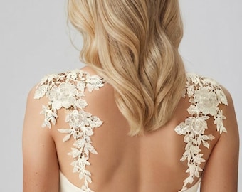 May include: An ivory-coloured sleeveless dress featuring intricate cream-coloured floral lace appliques on the shoulders and upper back. The garment includes a central vertical zip closure on the back and displays the text Orphelia in the corner.
