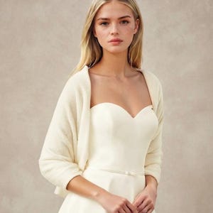 May include: A cream-colored knit bolero shrug worn over a strapless ivory dress with a sweetheart neckline and a thin waist belt. The bolero features long sleeves and a soft, textured knit fabric that drapes over the shoulders and upper arms.
