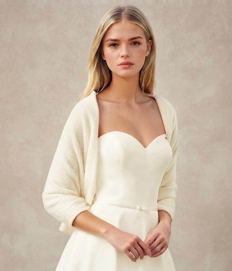 May include: A cream-colored knit bolero shrug worn over a strapless ivory dress with a sweetheart neckline and a thin waist belt. The bolero features long sleeves and a soft, textured knit fabric that drapes over the shoulders and upper arms.