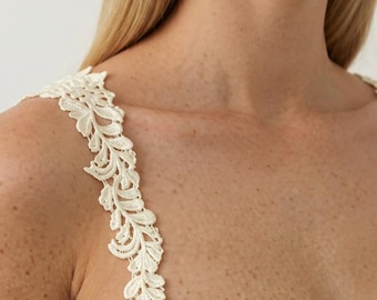 Peut inclure: Une robe blanc cassé avec une encolure en cœur et une large bretelle ornée d'une broderie en dentelle florale couleur crème. Le motif en dentelle se compose de feuilles et de vignes détaillées s'étendant le long de la bretelle.