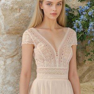 May include: A cream dress with a lace bodice and a flowing skirt. The dress has a V-neck and short sleeves. The model is standing in front of a stone wall with blue flowers.