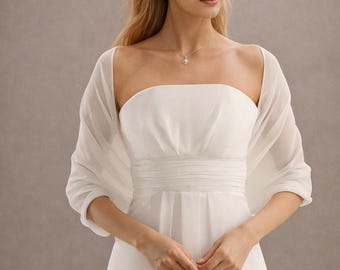 May include: A white strapless dress featuring a gathered bodice with a horizontal pleated waistband and a flowing skirt. A sheer, lightweight white shawl is draped over the shoulders and arms, complementing the clean, solid white fabric of the dress.