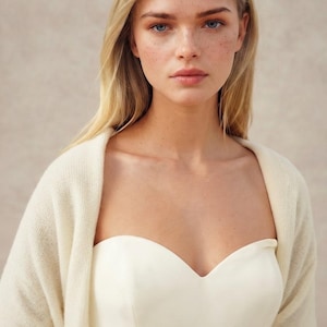 May include: A strapless cream-colored bodice paired with a soft, long-sleeved cream-colored knit cardigan. The garment features a sweetheart neckline and a smooth, solid texture, providing a layered look with a cohesive, neutral color palette.