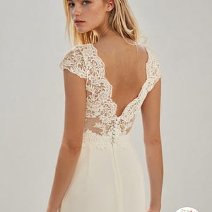May include: A floor-length ivory dress featuring a deep V-back and cap sleeves with intricate floral lace detailing. The bodice includes a sheer lace overlay, and the back is secured with a vertical row of small fabric-covered buttons.