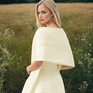 May include: A cream-colored, textured knit shawl draped over the shoulders, paired with a matching cream-colored sleeveless dress featuring a thin waist belt. The garment includes a circular logo in the bottom right corner with the text ORPHELIA.