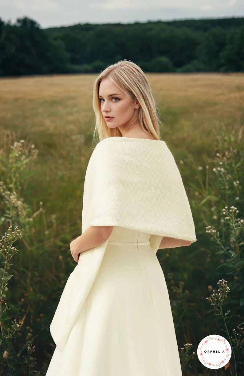 May include: A cream-colored, textured knit shawl draped over the shoulders, paired with a matching cream-colored sleeveless dress featuring a thin waist belt. The garment includes a circular logo in the bottom right corner with the text ORPHELIA.
