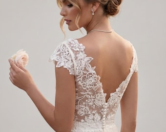 May include: A white floor-length gown featuring a sheer lace bodice with intricate floral embroidery, cap sleeves, and a deep V-shaped back. The bodice is accented with a vertical row of small fabric-covered buttons along the center back.