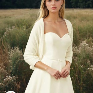 May include: An ivory strapless sweetheart neckline dress with a thin waist belt and a matching long-sleeved cropped cardigan. The dress features a fitted bodice and a full skirt. A circular logo with the text ORPHELIA is in the bottom corner.