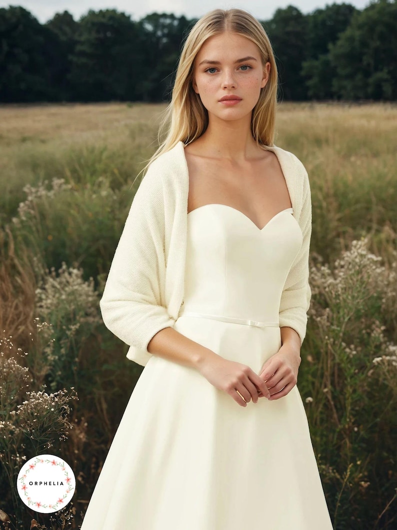 May include: An ivory strapless sweetheart neckline dress with a thin waist belt and a matching long-sleeved cropped cardigan. The dress features a fitted bodice and a full skirt. A circular logo with the text ORPHELIA is in the bottom corner.