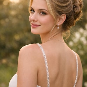 May include: A white sleeveless gown featuring thin straps adorned with clear sequins and beads. The back of the dress includes a vertical row of small white fabric-covered buttons. A delicate silver chain necklace and a single pearl drop earring are visible.