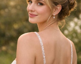 May include: A white sleeveless gown featuring thin straps adorned with clear sequins and beads. The back of the dress includes a vertical row of small white fabric-covered buttons. A delicate silver chain necklace and a single pearl drop earring are visible.