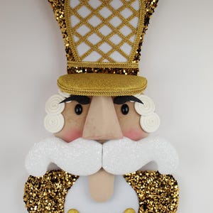 May include: A decorative nutcracker face with a gold sequined hat and collar. The hat features a white diamond pattern. The face has a light skin tone, black eyes, and a white, glittery mustache. Gold buttons adorn the white chest.