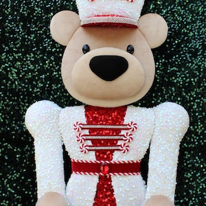 May include: A plush bear nutcracker decoration featuring a tan face with black eyes and nose. The bear wears a white sequined hat and jacket with red sequined accents, red velvet trim, small peppermint candy embellishments, and a central red gem.