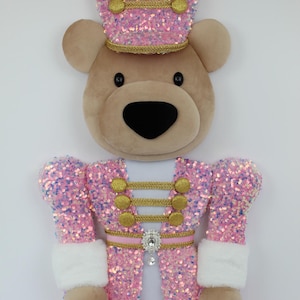 May include: A plush bear dressed as a nutcracker, featuring a pink sequined jacket and matching hat with gold trim and buttons. The bear has a tan face, black eyes, and a black nose. A white faux fur cuff accents the sleeves, and a rhinestone brooch.