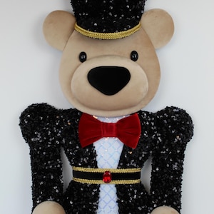 May include: A decorative wall hanging shaped like a bear dressed as a nutcracker soldier. The bear wears a black sequined hat with gold trim and red accents, a red velvet bow tie, and a black sequined jacket with a gold belt and red gem.