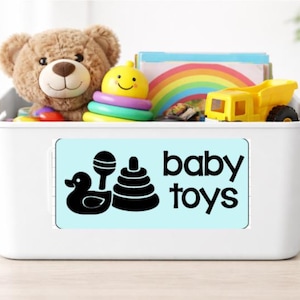May include: A white plastic bin filled with baby toys, including a teddy bear, stacking rings, a toy truck, and a rainbow-themed book. The bin has a light blue label that reads "baby toys" with illustrations.