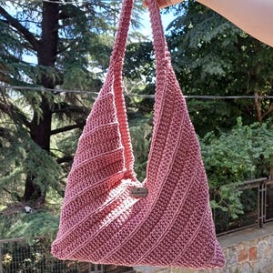 May include: A dusty rose crochet hobo bag featuring a diagonal ribbed texture and a single integrated shoulder strap. A small rectangular metal tag with the word handmade is attached to the center front of the bag, which has a triangular shape.