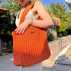 May include: A bright orange crochet tote bag featuring a vertical ribbed texture and a rectangular silver-toned metal brand tag attached to the upper front. The bag has a wide shoulder strap and is constructed from a thick, woven yarn material.