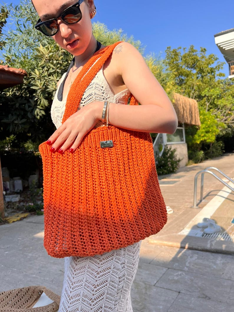 May include: A bright orange crochet tote bag featuring a vertical ribbed texture and a rectangular silver-toned metal brand tag attached to the upper front. The bag has a wide shoulder strap and is constructed from a thick, woven yarn material.