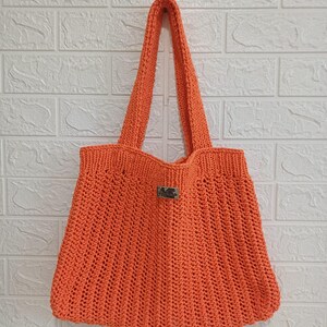 May include: A bright orange crochet tote bag featuring a textured ribbed stitch pattern throughout the body. The bag includes two long shoulder straps and a small rectangular silver-toned metal brand tag centered on the front upper section.
