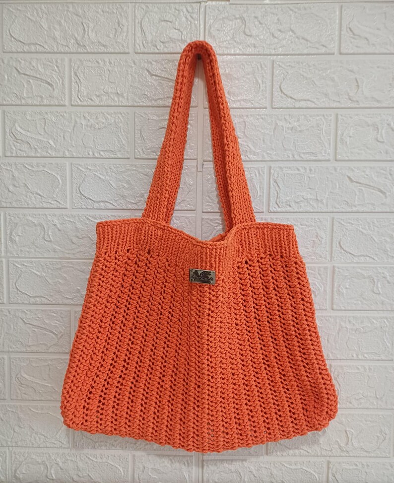 May include: A bright orange crochet tote bag featuring a textured ribbed stitch pattern throughout the body. The bag includes two long shoulder straps and a small rectangular silver-toned metal brand tag centered on the front upper section.
