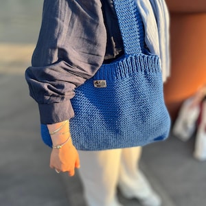 Könnte beinhalten: Eine leuchtend blaue, gestrickte Tragetasche mit einer texturierten kraus rechts gestrickten Struktur, einem breiten Schulterriemen und einem kleinen, rechteckigen Metall-Markenetikett am oberen vorderen Rand der Tasche.