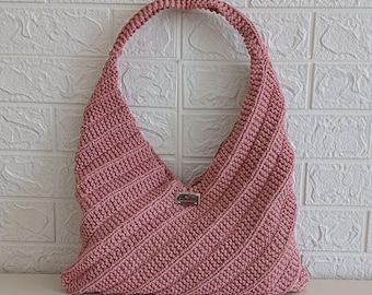 May include: A dusty rose crochet hobo bag featuring a diagonal ribbed texture and a single integrated shoulder strap. A small rectangular metallic brand tag is centered on the front of the bag, which is constructed from thick yarn stitches.