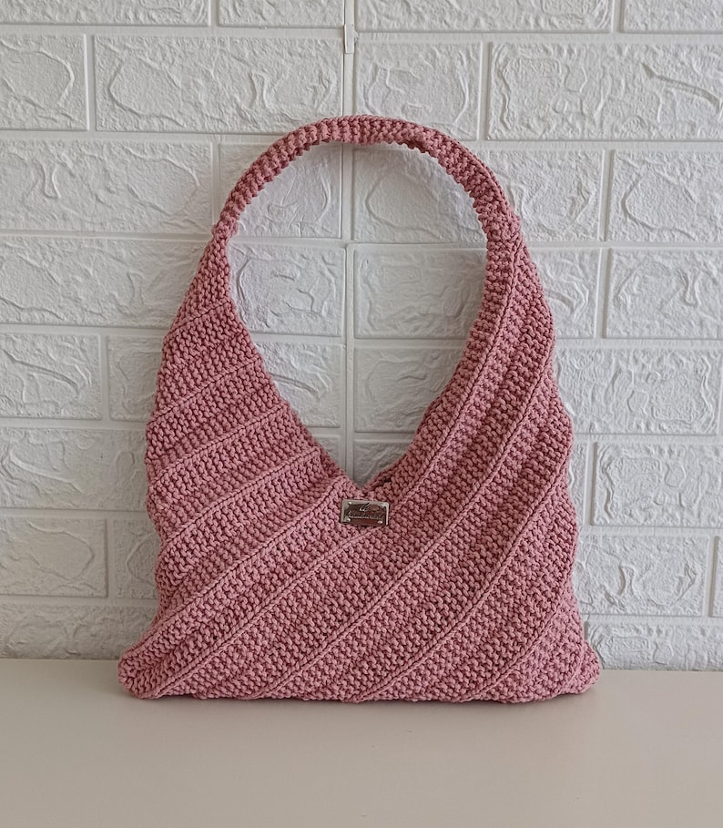 Pu&ograve; includere: Una borsa hobo all'uncinetto color rosa antico caratterizzata da una trama a coste diagonali su tutto il corpo. La borsa presenta un unico spallaccio integrato e una piccola etichetta metallica rettangolare centrata sulla parte anteriore.