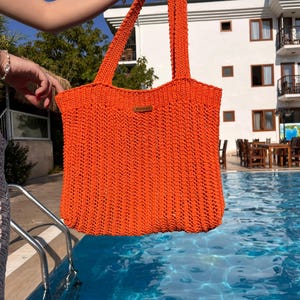 May include: A bright orange crochet tote bag featuring a vertical ribbed stitch pattern and two long shoulder straps. A small rectangular brown leather-like tag is attached to the upper center of the bag, displaying the embossed text Handmade.