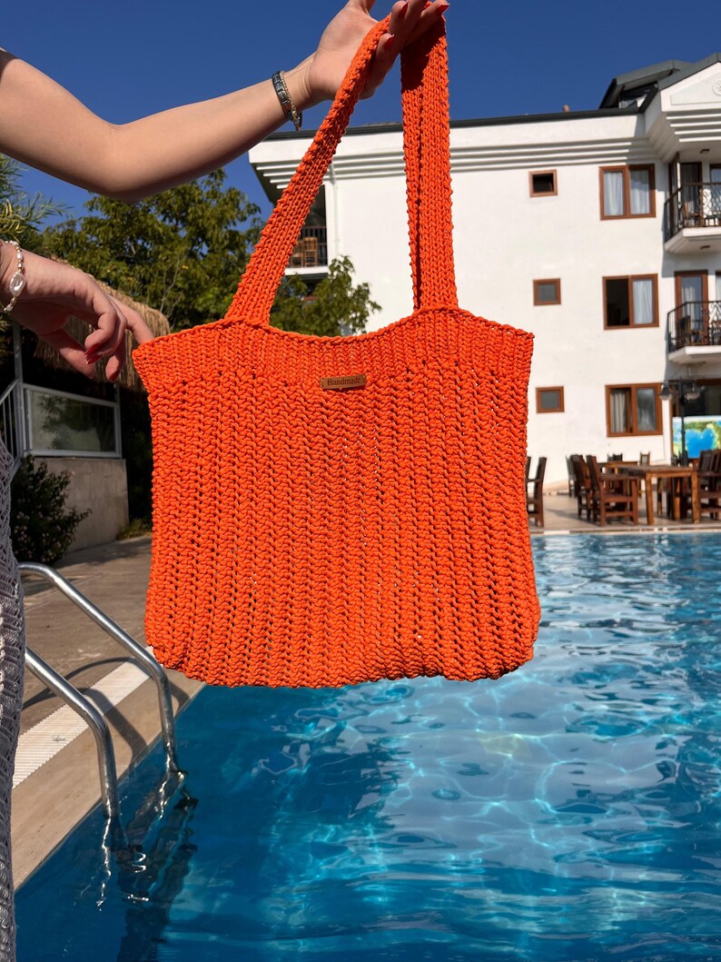 May include: A bright orange crochet tote bag featuring a vertical ribbed stitch pattern and two long shoulder straps. A small rectangular brown leather-like tag is attached to the upper center of the bag, displaying the embossed text Handmade.