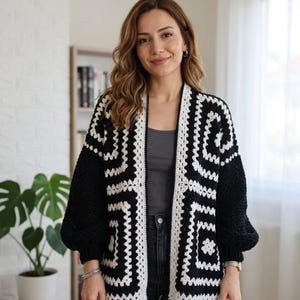 May include: A black and white crochet cardigan featuring a geometric granny square pattern. The garment has an open front design, long sleeves with ribbed cuffs, and a contrasting white border that frames the edges of the black and white squares.