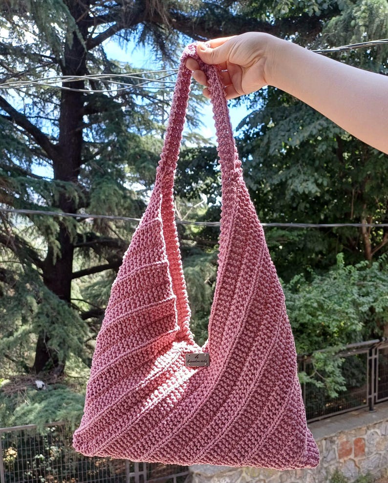 Pu&ograve; includere: Una borsa hobo all'uncinetto color rosa polvere caratterizzata da una trama a coste diagonali e un unico manico a spalla integrato. Una piccola targhetta metallica rettangolare con la parola handmade &egrave; fissata al centro della parte anteriore della borsa, che ha una forma triangolare.