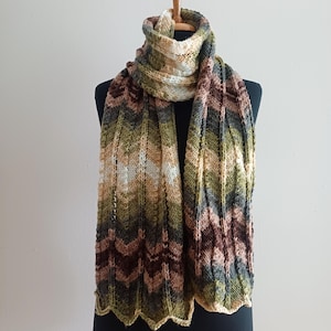 Könnte beinhalten: Ein gestrickter Schal in Braun-, Grün- und Beigetönen, präsentiert auf einer schwarzen Schaufensterpuppe. Der Schal hat ein Zickzackmuster und einen gewellten Rand. Der Schal ist etwa 178 cm lang.