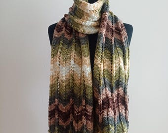 May include: A long, hand-knitted scarf featuring a repeating chevron or ripple pattern. The yarn displays a variegated color palette of earthy tones, including shades of forest green, dark brown, tan, cream, and muted grey, arranged in horizontal stripes.