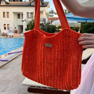 May include: A bright orange crochet tote bag featuring a vertical ribbed stitch pattern and two long shoulder straps. A small rectangular metal tag with the text "le handmade" is attached to the center front of the bag.